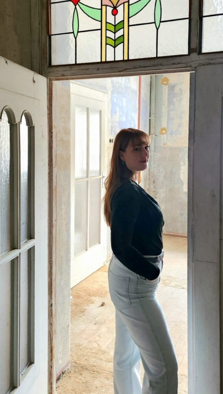 Die Galeristin steht in einem hellen Raum mit Flügeltür und Buntglasfenster im Jugendstil. Die Tür verbindet die Galerieräume miteinander.